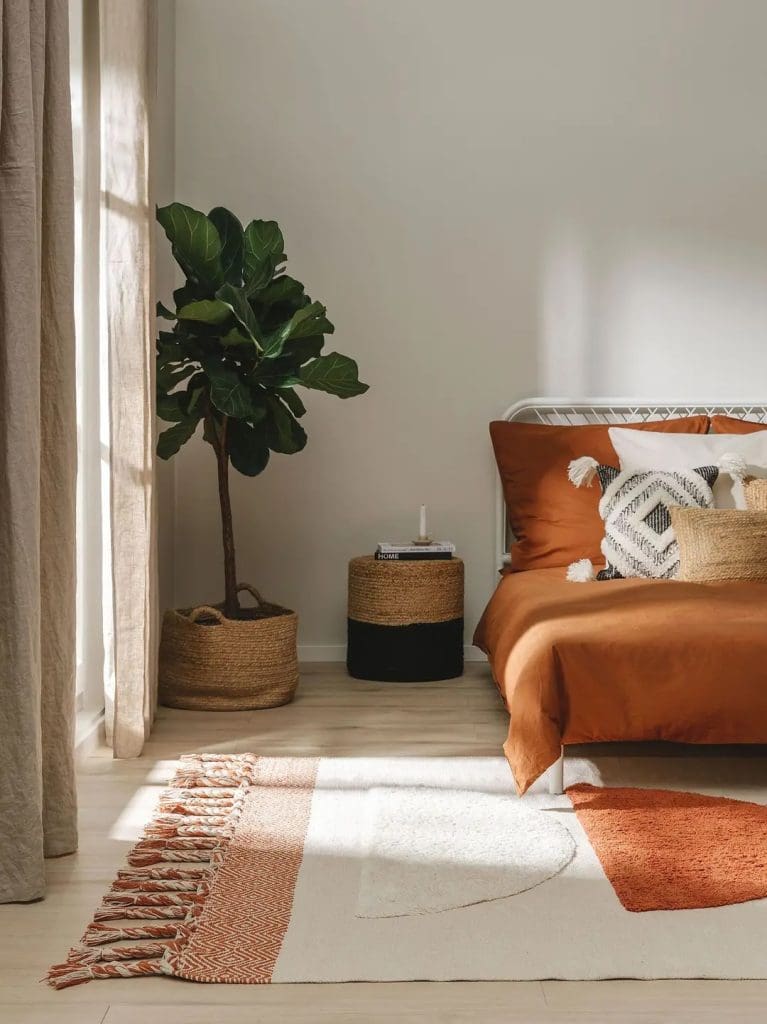 Light and relaxing bedroom with tall fig tree in the corner, terracotta bedding and boho patterned cushions, as well as a off-white and terracotta tasselled rug